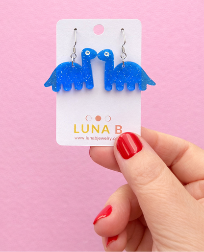 Blue dinosaur-shaped earrings on a 'Luna B' branded card against a pink background.