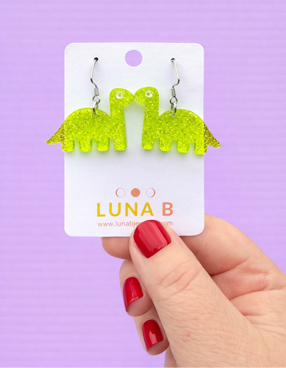 Green dinosaur-shaped earrings on a Luna B card held by a hand with red nails against a purple background