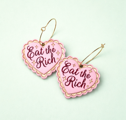 Pink earrings with 'Eat the Rich' text on a light green background