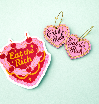 Heart-shaped earrings with 'Eat the Rich' text on a light green background