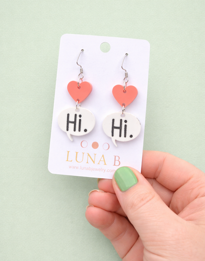 Heart-shaped earrings on a Luna B card held by a hand with green nail polish against a light green background.