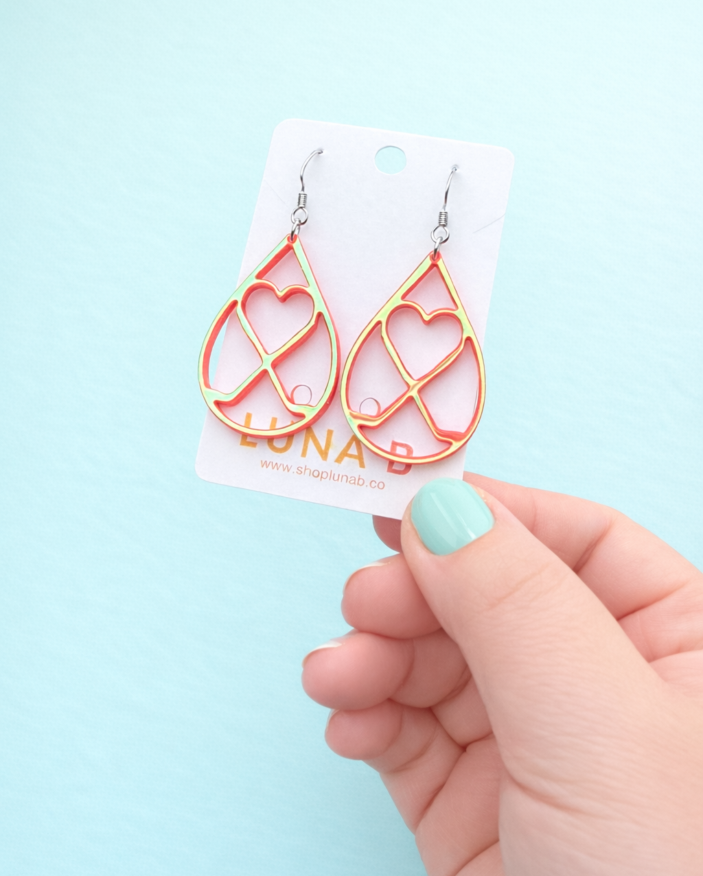 Iridescent pink with a golden hew heart-shaped earrings on a white card held by a hand against a light blue background