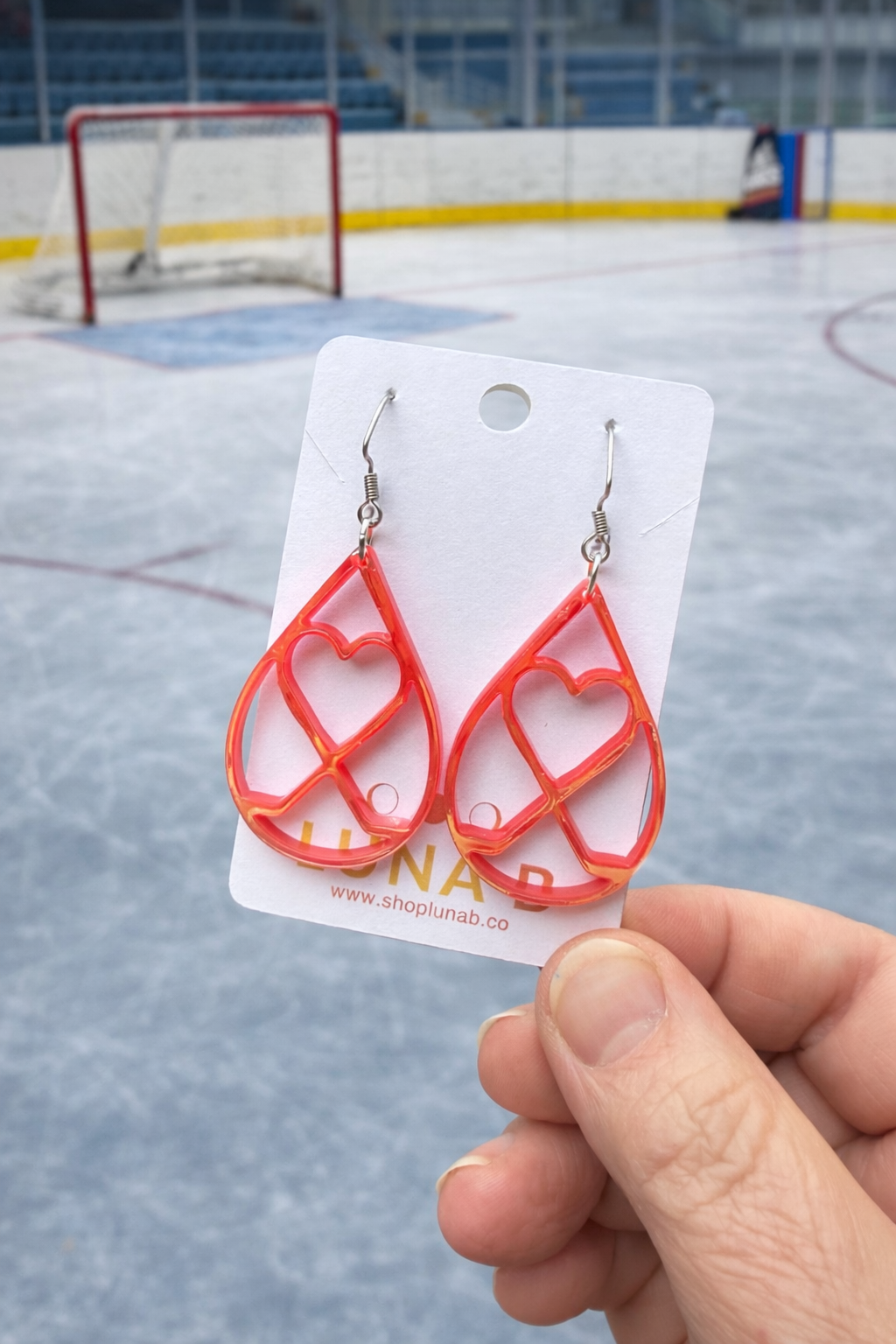 Red heart-shaped earrings on a white card held in front of an ice rink.