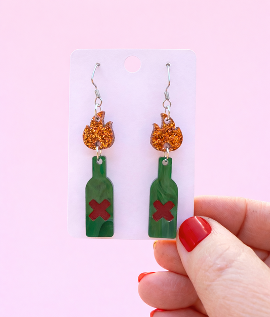 Green bottle-shaped earrings with orange flames held by a hand against a pink background