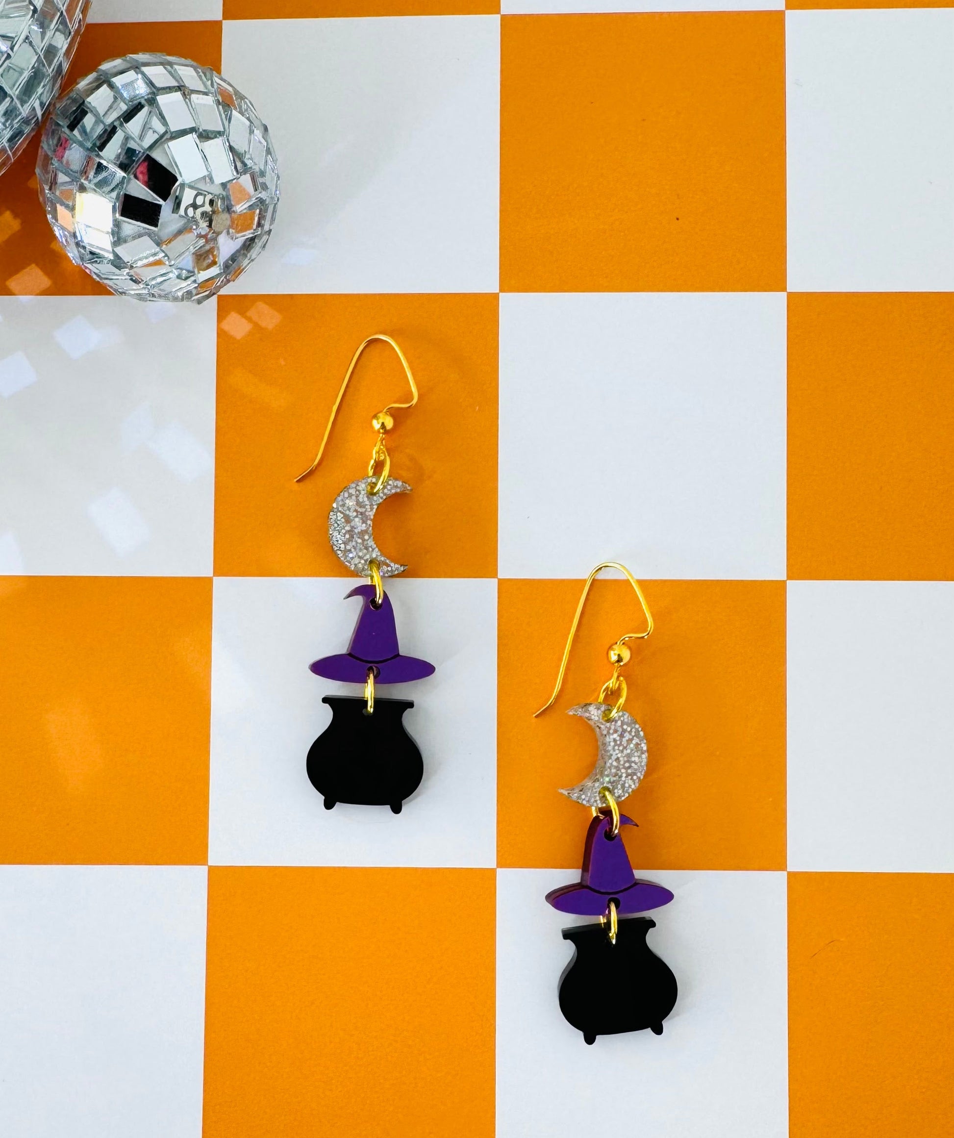 Pair of earrings with black cat and witch hat design on a checkered orange and white background.