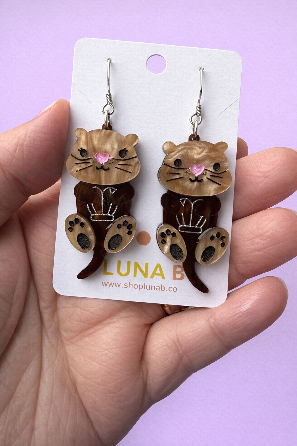 Otter-shaped earrings on a Luna B branded card held by a hand against a purple background