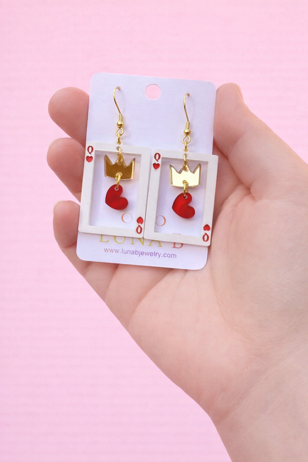 Hand holding Luna B Queen of Hearts earrings featuring red heart charms and gold queen details on stainless steel gold-plated ear wires, displayed on a branded card against a soft pastel rainbow background.