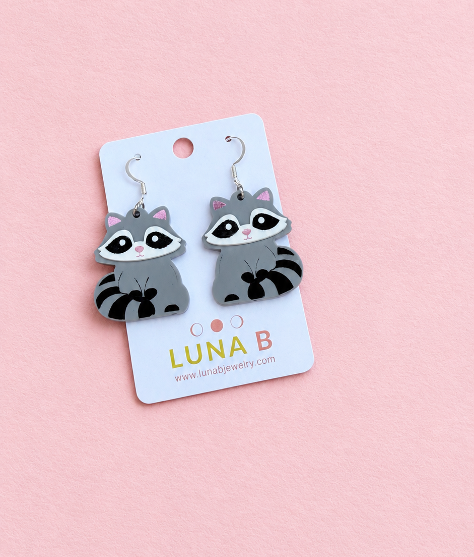 A pair of raccoon-shaped dangle earrings on a white display card, photographed on a soft pink background. The card is branded LUNA B.