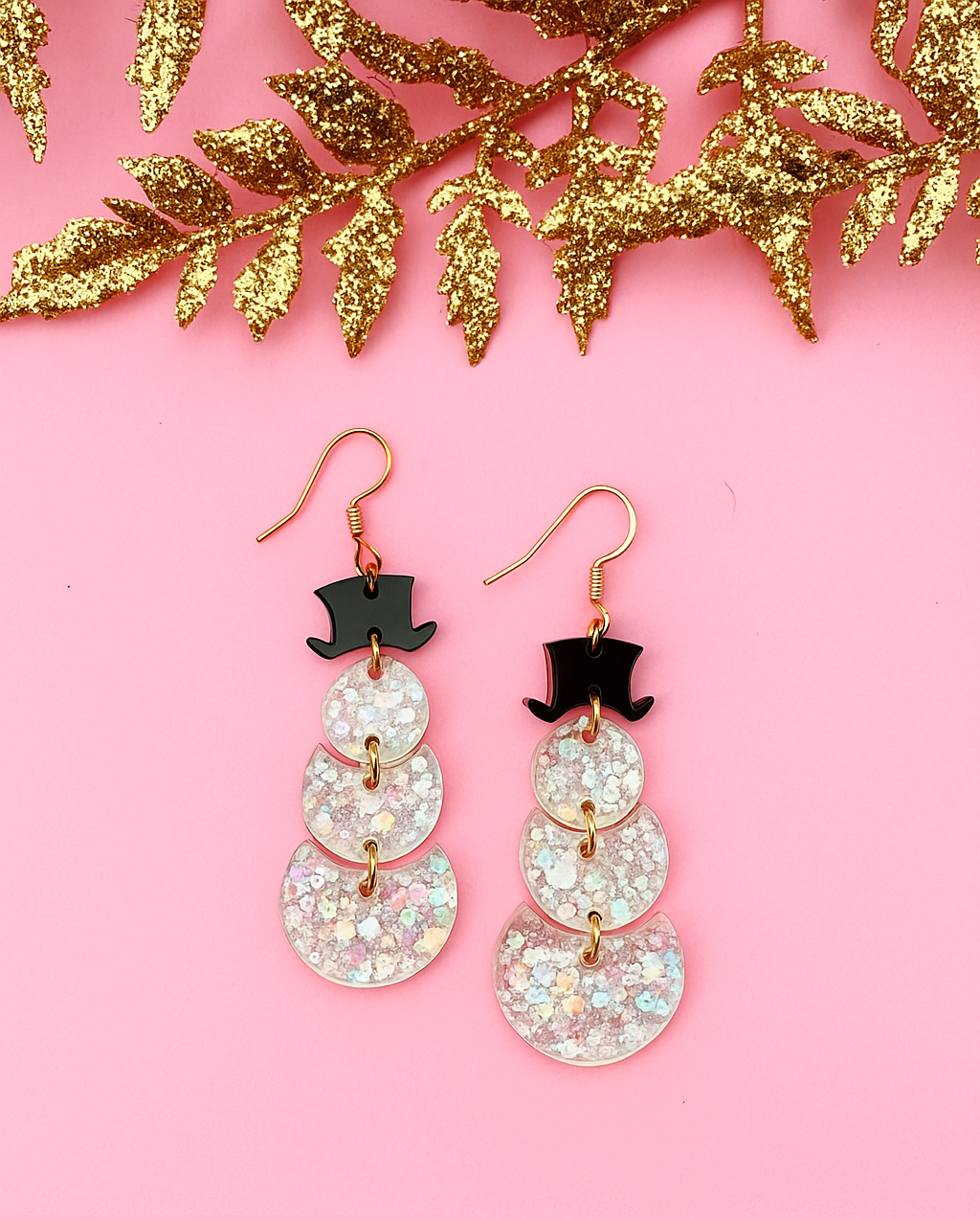 Sparkly snowman earrings with top hats on a pink background with gold leaves.