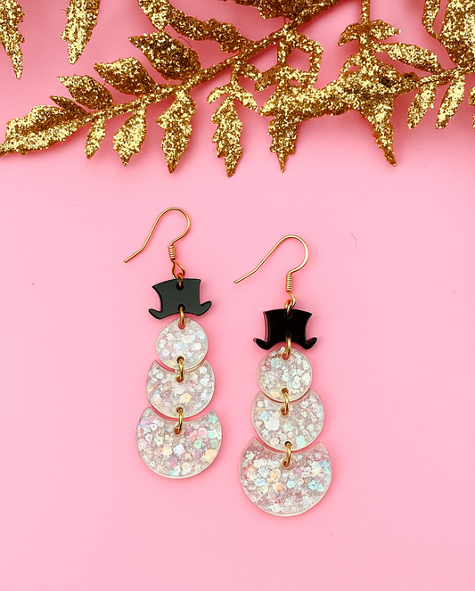 Sparkly snowman earrings with top hats on a pink background with gold leaves.