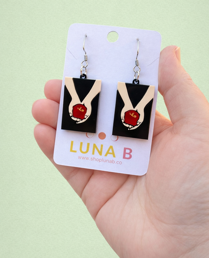 Layered acrylic earrings featuring pale hands holding a red apple on stainless steel hooks, displayed on a Luna B Jewelry card.