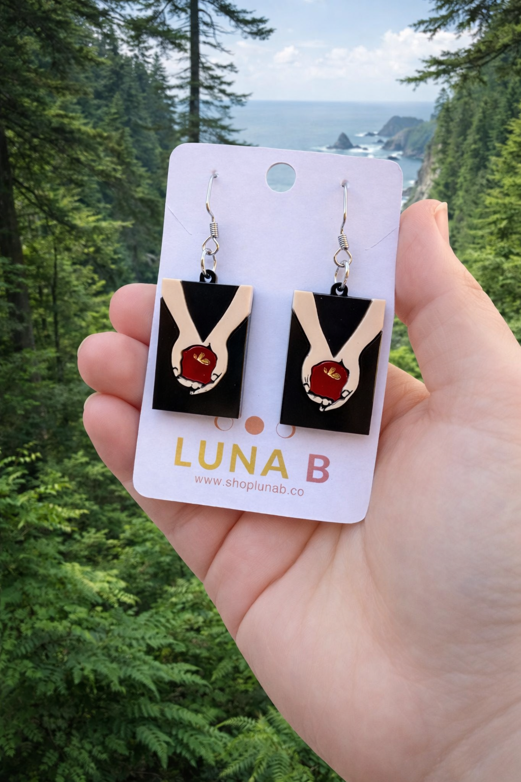 Layered acrylic earrings featuring pale hands holding a red apple on stainless steel hooks, displayed on a Luna B Jewelry card.