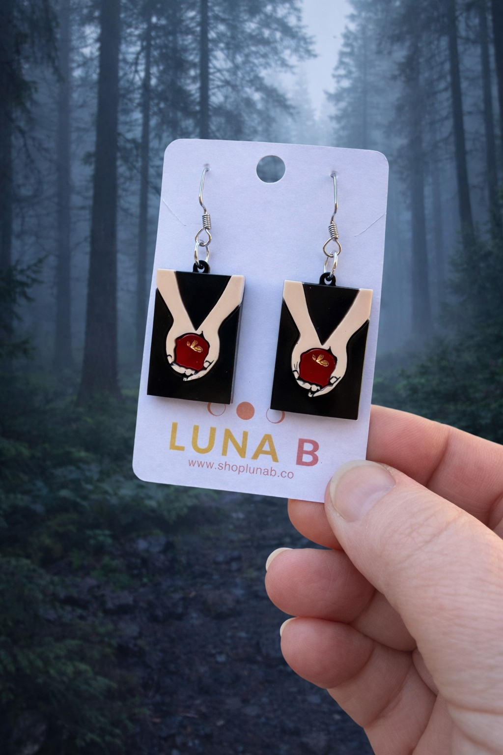 Layered acrylic earrings featuring pale hands holding a red apple on stainless steel hooks, displayed on a Luna B Jewelry card.