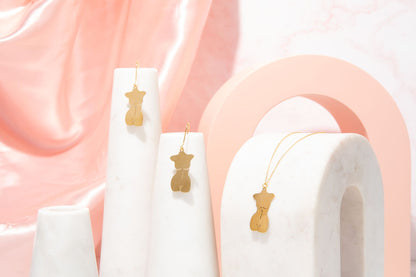 Gold feminine body earrings displayed on white cylindrical stands against a soft pink background