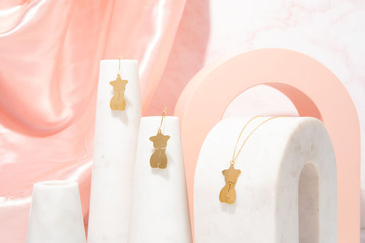 Gold feminine body earrings displayed on white cylindrical stands against a soft pink background